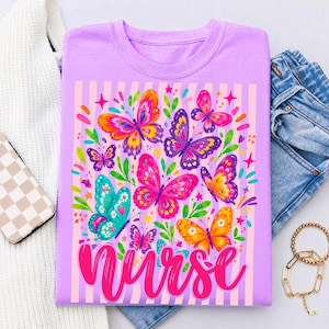 May include: A light purple t-shirt featuring a graphic print of colorful butterflies in shades of pink, orange, blue, and purple, accented with green leaves and small stars. The word nurse is printed in a bold, cursive pink font at the bottom.
