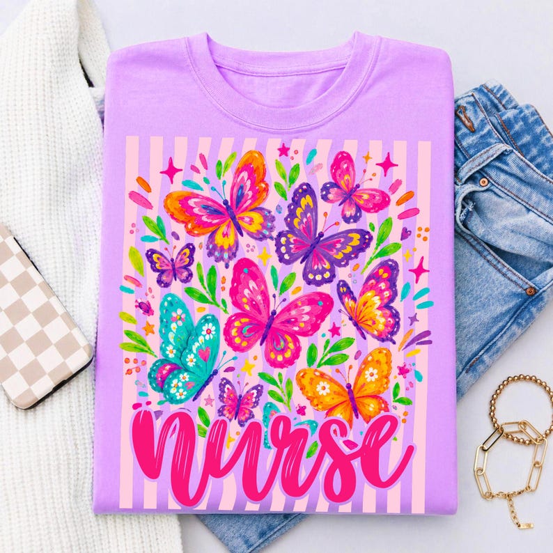 May include: A light purple t-shirt featuring a graphic print of colorful butterflies in shades of pink, orange, blue, and purple, accented with green leaves and small stars. The word nurse is printed in a bold, cursive pink font at the bottom.