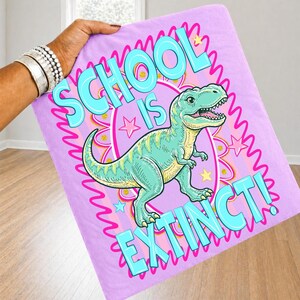 Op de afbeelding: Een lichtpaarse deken met een cartoon Tyrannosaurus Rex illustratie. De deken heeft de tekst "SCHOOL IS EXTINCT!" in turquoise en roze. De dinosaurus is lichtgroen en geel, met een roze en turquoise rand.