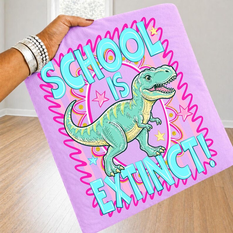 Op de afbeelding: Een lichtpaarse deken met een cartoon Tyrannosaurus Rex illustratie. De deken heeft de tekst "SCHOOL IS EXTINCT!" in turquoise en roze. De dinosaurus is lichtgroen en geel, met een roze en turquoise rand.