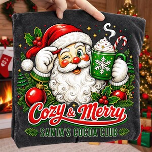 K&ouml;nnte beinhalten: Ein dunkelgraues T-Shirt mit einer Grafik vom zwinkernden Weihnachtsmann, der eine gr&uuml;ne Tasse mit wei&szlig;em Schneeflockenmuster und eine Zuckerstange h&auml;lt. Der Text lautet Cozy & Merry SANTA'S COCOA CLUB, umgeben von Stechpalmen, Tannen und roten Kugeln.