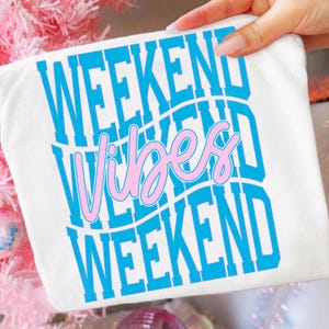 K&ouml;nnte beinhalten: Wei&szlig;es T-Shirt mit den Worten "WEEKEND VIBES WEEKEND" in blauem und rosa Design. Der Text ist vertikal gestapelt, mit dem Wort "Vibes" in einer Schreibschrift. Das Shirt wird von einer Hand gehalten.