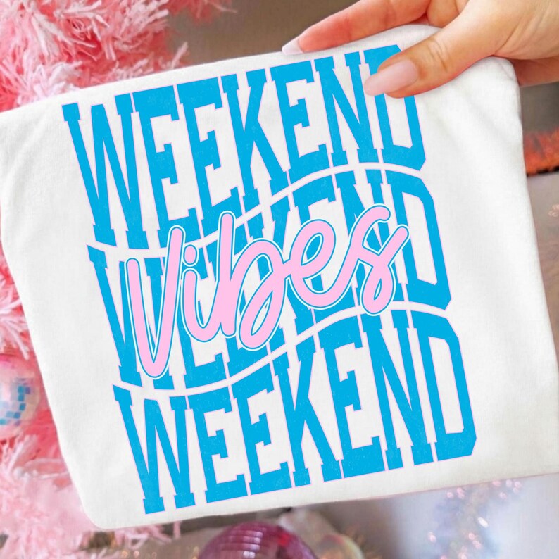 K&ouml;nnte beinhalten: Wei&szlig;es T-Shirt mit den Worten "WEEKEND VIBES WEEKEND" in blauem und rosa Design. Der Text ist vertikal gestapelt, mit dem Wort "Vibes" in einer Schreibschrift. Das Shirt wird von einer Hand gehalten.