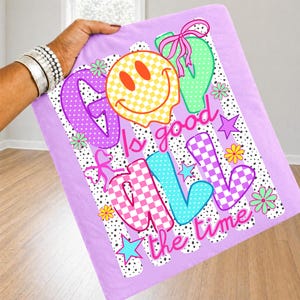 Op de afbeelding: Een lichtpaars t-shirt met een grafische print met de tekst God is good all the time. Het ontwerp bevat een geel geblokt smileygezicht, roze strikken, kleurrijke bloemen, sterren en een achtergrond van zwarte stippen en verticale strepen.