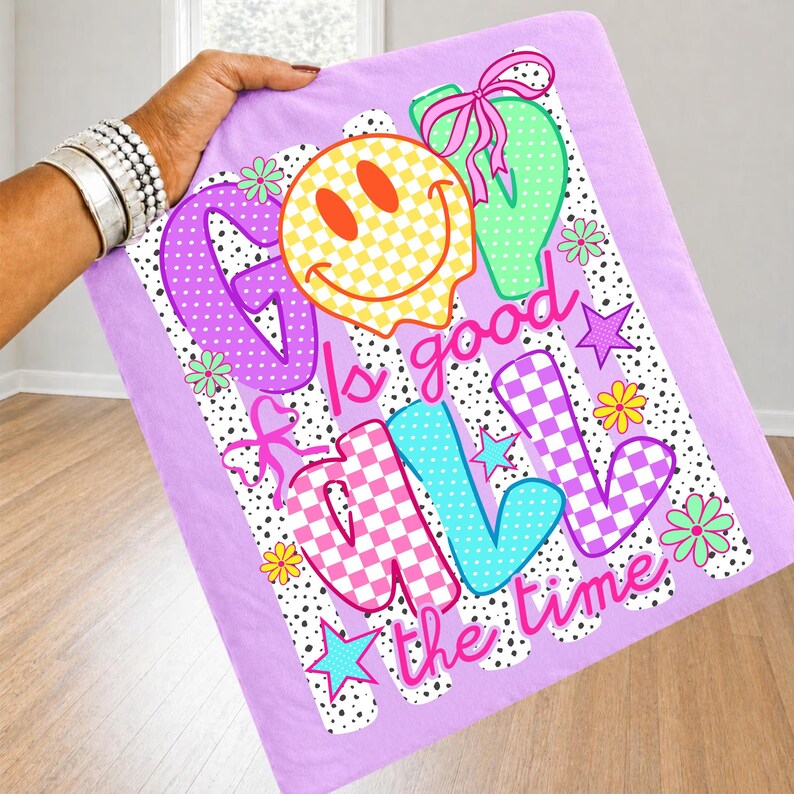 Op de afbeelding: Een lichtpaars t-shirt met een grafische print met de tekst God is good all the time. Het ontwerp bevat een geel geblokt smileygezicht, roze strikken, kleurrijke bloemen, sterren en een achtergrond van zwarte stippen en verticale strepen.