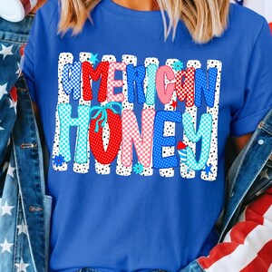 May include: A royal blue short-sleeved t-shirt featuring a graphic print with the text AMERICAN HONEY. The letters are filled with red, white, and blue polka dot and checkered patterns, set against a background of vertical black and white stripes.