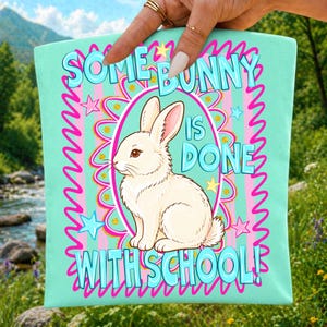 Może przedstawiać: Jasnoniebieska torba z napisem "SOME BUNNY IS DONE WITH SCHOOL!" i ilustracją białego kr&oacute;lika. Projekt zawiera r&oacute;żowe i złote akcenty, gwiazdki i ozdobną obw&oacute;dkę. Torba jest trzymana przez dłoń.