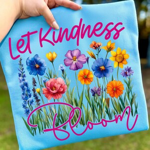 May include: A light blue tote bag featuring the words "Let Kindness Bloom" in pink script. The design includes a colorful floral arrangement with blue, purple, yellow, and orange flowers, and green grass.