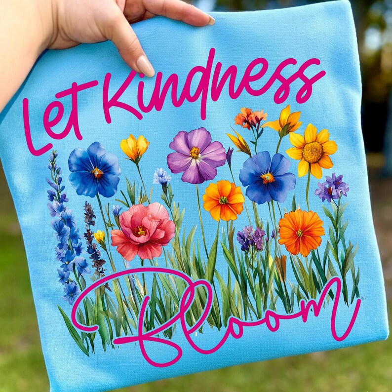 May include: A light blue tote bag featuring the words "Let Kindness Bloom" in pink script. The design includes a colorful floral arrangement with blue, purple, yellow, and orange flowers, and green grass.
