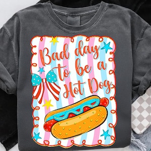 May include: Dark gray sweatshirt with a colorful graphic that says "Bad day to be a Hot Dog." The graphic includes a hot dog, stars, and a bow with red, white, and blue stripes.