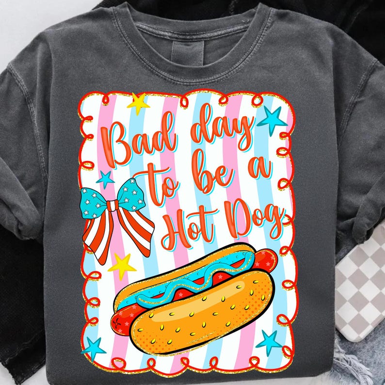 May include: Dark gray sweatshirt with a colorful graphic that says "Bad day to be a Hot Dog." The graphic includes a hot dog, stars, and a bow with red, white, and blue stripes.
