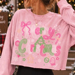 May include: A light pink long-sleeve cropped sweatshirt featuring the text Merry Christmas in a wavy font. The design includes a pink bow, a green Christmas tree, a candy cane, a gingerbread man, a mitten, holly berries, and white snowflakes.
