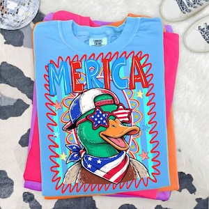 May include: A light blue t-shirt with the word "MERICA" in red and blue lettering. The shirt features a cartoon duck wearing sunglasses, a hat, and a bandana, all in patriotic colors. The shirt is folded on top of other colored shirts.