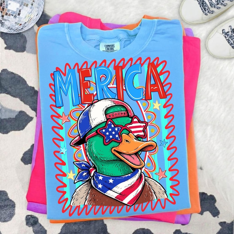 May include: A light blue t-shirt with the word "MERICA" in red and blue lettering. The shirt features a cartoon duck wearing sunglasses, a hat, and a bandana, all in patriotic colors. The shirt is folded on top of other colored shirts.
