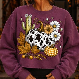 Puede incluir: Una sudadera de manga larga color ciruela con un estampado gr&aacute;fico de una calabaza con manchas de vaca, una calabaza peque&ntilde;a de cuadros amarillos, un cactus verde con flores blancas, hojas de oto&ntilde;o, bellotas, espigas de trigo y margaritas.