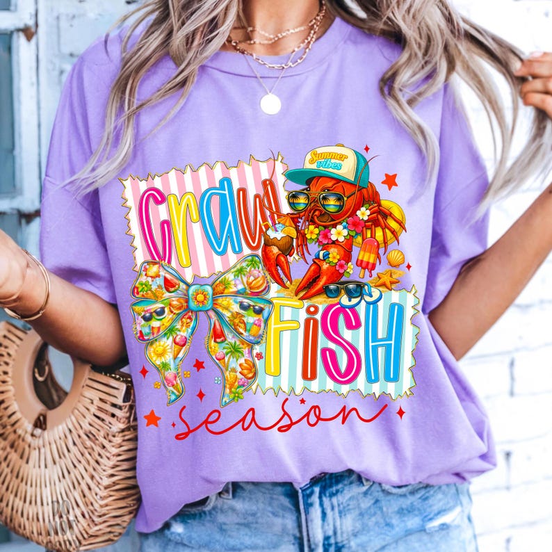 May include: A lilac t-shirt with a vibrant graphic reading "Crawfish Season". The design includes a cartoon crawfish in sunglasses and a cap, a bow, and other summery illustrations.