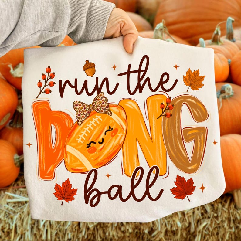 Puede incluir: Una camiseta blanca con el texto 'run the ball' en letra cursiva marr&oacute;n. La letra 'O' es un bal&oacute;n de f&uacute;tbol americano naranja con un lazo de estampado de leopardo y una cara sonriente. Incluye hojas de arce naranjas, rojas y bayas.