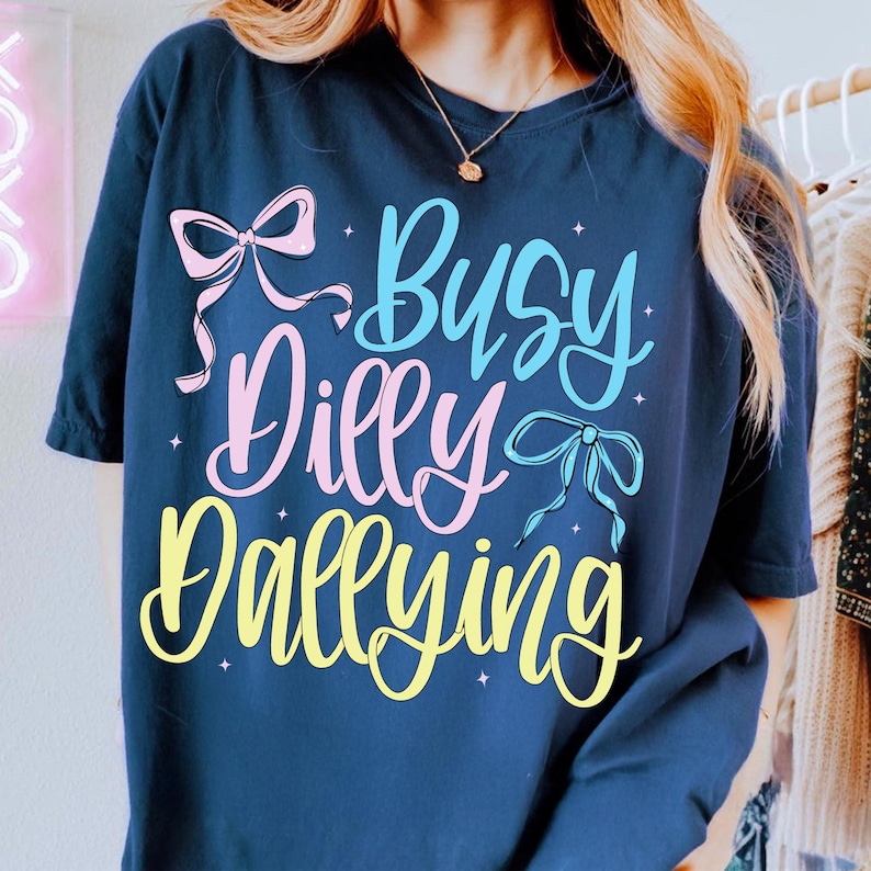 Puede incluir: Camiseta azul marino con la frase "Busy Dilly Dallying" en una fuente colorida y juguetona. El dise&ntilde;o incluye lazos rosas y azules. La camiseta tiene un corte holgado.