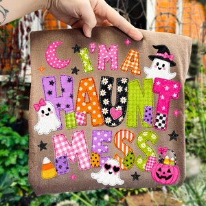 May include: A brown t-shirt featuring the colorful text "I'M A HAUNT MESS" in various patterns like polka dots, stripes, and florals. The design includes three white ghost illustrations, a pink jack-o'-lantern bucket, and candy corn.