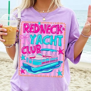 May include: A lilac-colored t-shirt with a graphic that reads "Redneck Yacht Club." The graphic features a pink and blue pontoon boat, stars, and a bow. The shirt is a relaxed fit.