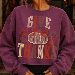 Puede incluir: Una sudadera de manga larga color ciruela con el texto GIVE THANKS en una fuente superpuesta de varios tonos. Centrado entre las palabras hay un gr&aacute;fico de una calabaza con un lazo, rodeado por varios contornos de hojas de oto&ntilde;o en naranja y marr&oacute;n.
