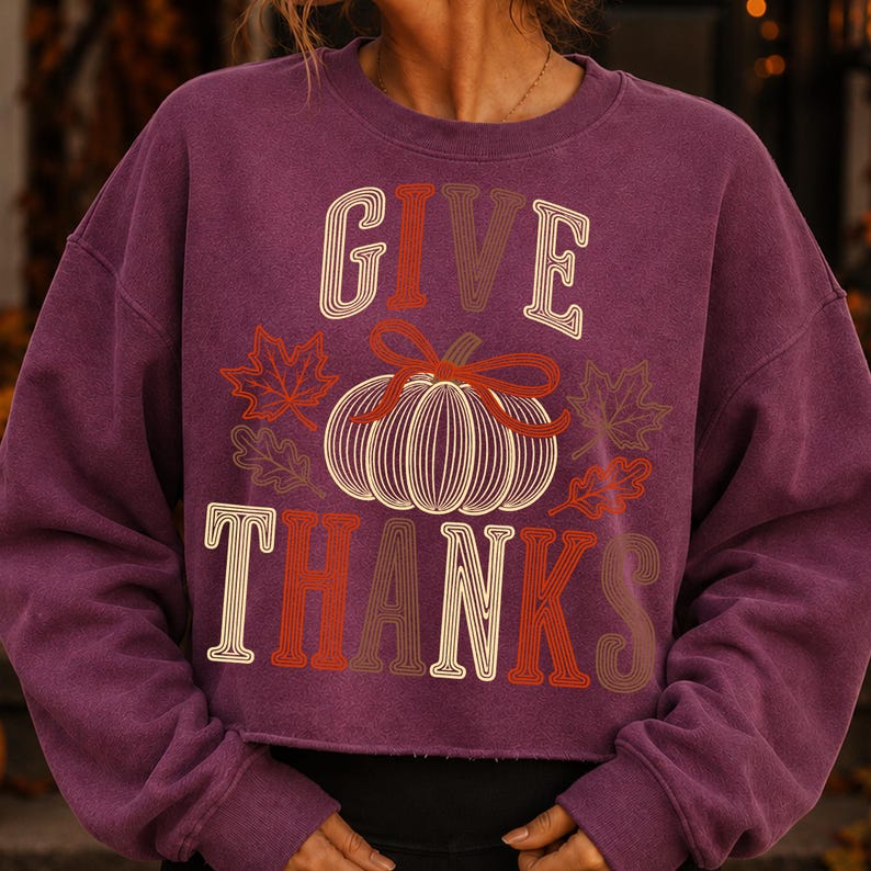 Puede incluir: Una sudadera de manga larga color ciruela con el texto GIVE THANKS en una fuente superpuesta de varios tonos. Centrado entre las palabras hay un gr&aacute;fico de una calabaza con un lazo, rodeado por varios contornos de hojas de oto&ntilde;o en naranja y marr&oacute;n.