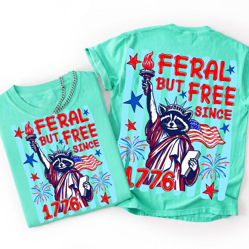 May include: Two turquoise t-shirts with a graphic of a raccoon dressed as the Statue of Liberty, holding a torch and a tablet, with an American flag. The text reads "FERAL BUT FREE SINCE 1776". Stars and fireworks are also featured.
