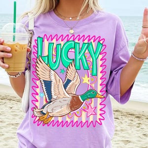May include: A lilac t-shirt featuring a cartoon duck in flight, with the word "LUCKY" in green. The duck has brown, blue, and white feathers. The design includes stars and a pink wavy border.