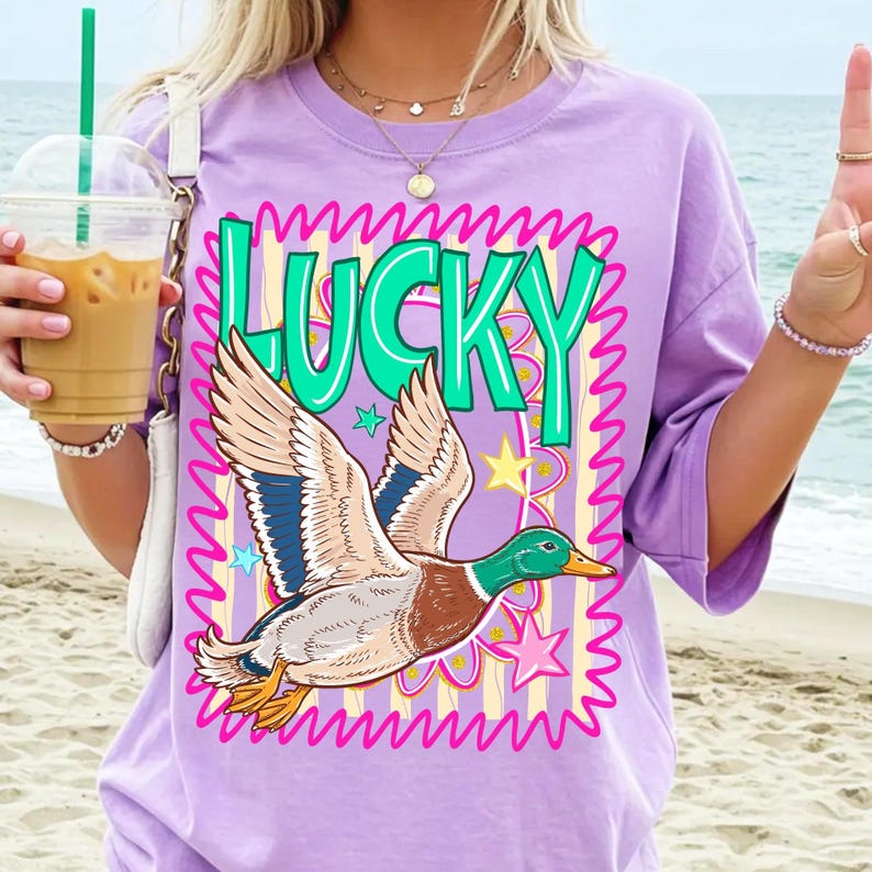 May include: A lilac t-shirt featuring a cartoon duck in flight, with the word "LUCKY" in green. The duck has brown, blue, and white feathers. The design includes stars and a pink wavy border.