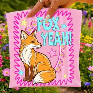 Op de afbeelding: Roze grafisch T-shirt met een cartoonvos in oranje en wit. De turquoise tekst luidt "FOX YEAH!", omlijst door sterren en een golvende roze rand. Het T-shirt wordt gepresenteerd tegen een achtergrond van kleurrijke bloemen.