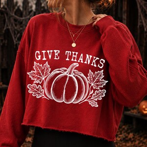 Puede incluir: Una sudadera roja de manga larga y corte corto con un estampado gr&aacute;fico blanco de una calabaza rodeada de hojas de arce. El texto "GIVE THANKS" est&aacute; impreso en blanco sobre la ilustraci&oacute;n de la calabaza. El dobladillo es de corte crudo.