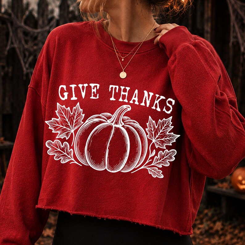 Puede incluir: Una sudadera roja de manga larga y corte corto con un estampado gr&aacute;fico blanco de una calabaza rodeada de hojas de arce. El texto "GIVE THANKS" est&aacute; impreso en blanco sobre la ilustraci&oacute;n de la calabaza. El dobladillo es de corte crudo.