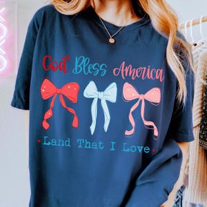 Może przedstawiać: Granatowa koszulka z napisem "God Bless America" w czerwonym i niebieskim kolorze. Poniżej tekstu znajdują się trzy kokardy w kolorze czerwonym, jasnoniebieskim i r&oacute;żowym. Pod kokardami znajduje się napis "Land That I Love".