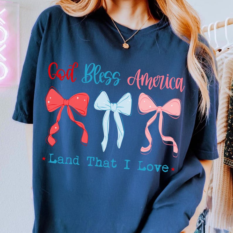 Może przedstawiać: Granatowa koszulka z napisem "God Bless America" w czerwonym i niebieskim kolorze. Poniżej tekstu znajdują się trzy kokardy w kolorze czerwonym, jasnoniebieskim i r&oacute;żowym. Pod kokardami znajduje się napis "Land That I Love".