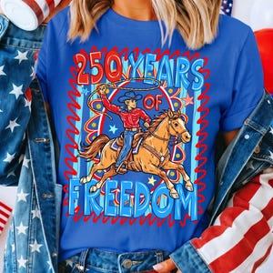 Puede incluir: Camiseta azul con un dise&ntilde;o de un vaquero a caballo con un lazo. El texto dice "250 YEARS OF FREEDOM" en rojo y azul. El dise&ntilde;o tiene un tema patri&oacute;tico.