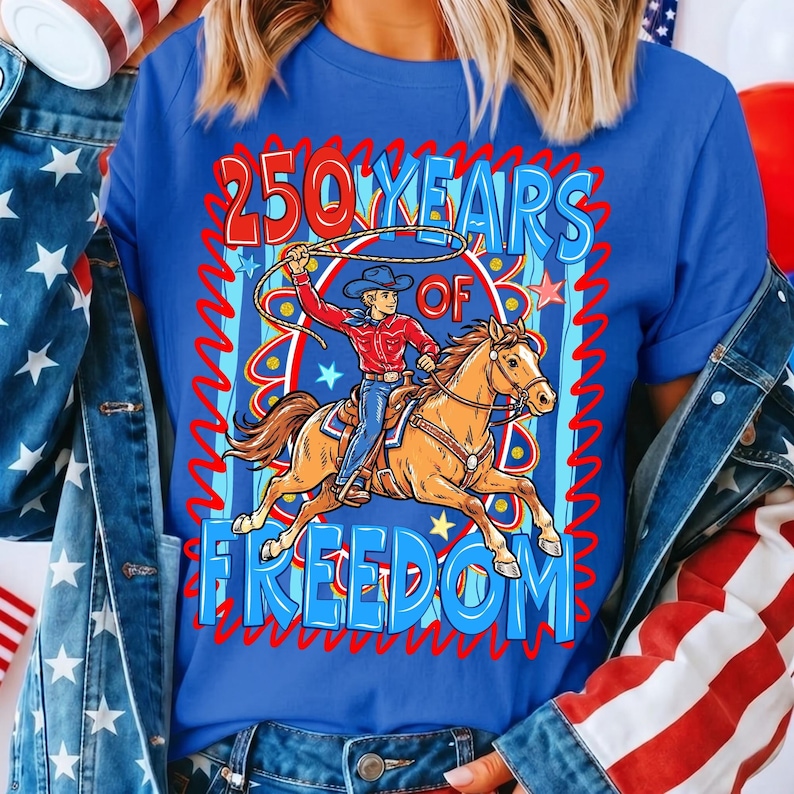 Puede incluir: Camiseta azul con un dise&ntilde;o de un vaquero a caballo con un lazo. El texto dice "250 YEARS OF FREEDOM" en rojo y azul. El dise&ntilde;o tiene un tema patri&oacute;tico.
