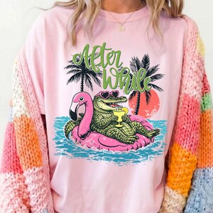 Pu&ograve; includere: T-shirt rosa chiaro con una grafica di un alligatore con occhiali da sole e un drink, sdraiato su un galleggiante a forma di fenicottero rosa. Il testo "After While" &egrave; sopra l'alligatore, con palme e un tramonto sullo sfondo.