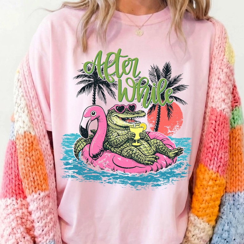 Pu&ograve; includere: T-shirt rosa chiaro con una grafica di un alligatore con occhiali da sole e un drink, sdraiato su un galleggiante a forma di fenicottero rosa. Il testo "After While" &egrave; sopra l'alligatore, con palme e un tramonto sullo sfondo.