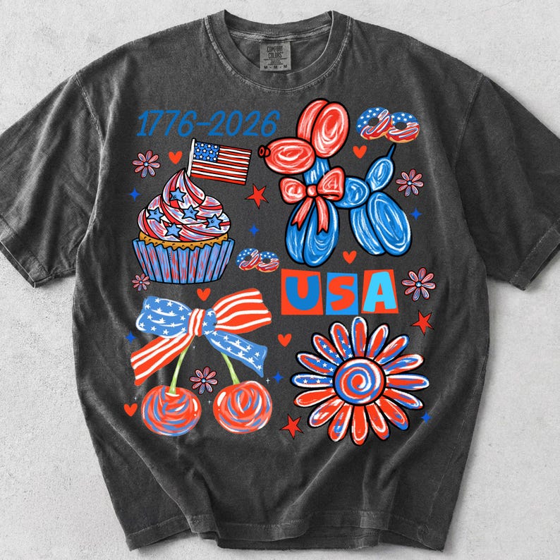 Pode incluir: T-shirt cinzenta escura com um design patri&oacute;tico. O gr&aacute;fico inclui um cupcake com uma bandeira, animais de bal&atilde;o, cerejas, uma flor e os textos "USA" e "1776-2026". O design utiliza as cores vermelho, branco e azul.