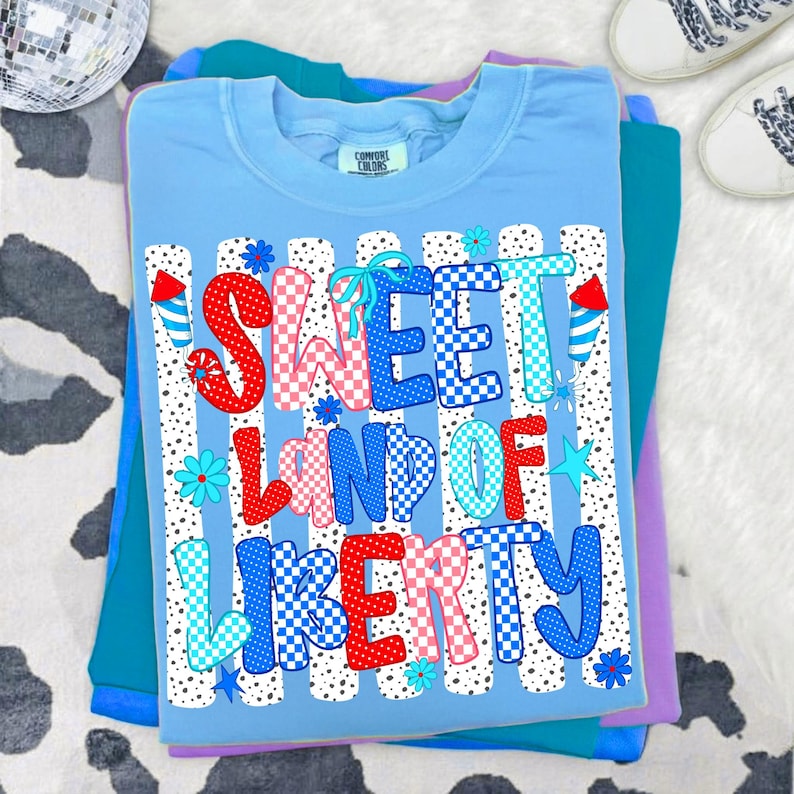 May include: A light blue short-sleeve t-shirt featuring the text SWEET LAND OF LIBERTY in red, white, and blue checkered patterns. The design includes small blue flowers, stars, and firework graphics against a background of vertical dotted lines.