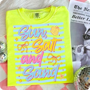 Peut inclure: Un t-shirt jaune n&eacute;on avec l'inscription "Sun, Salt, and Sand" dans une police color&eacute;e et ludique. Le motif comprend des coquillages, des n&oelig;uds et des &eacute;toiles de mer. Le t-shirt est &agrave; col rond et semble &ecirc;tre fait d'une mati&egrave;re douce.