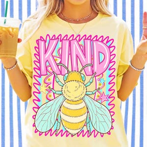 May include: Yellow t-shirt with a graphic of a bee and the word "KIND" in pink letters. The bee has yellow and black stripes, blue wings, and is surrounded by stars and wavy lines. The shirt is a casual style.