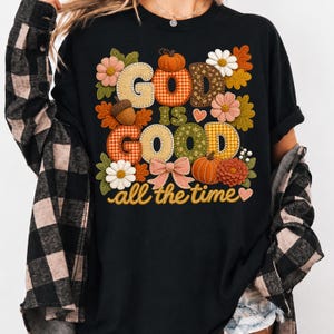 Pu&ograve; includere: Una t-shirt nera con una grafica che riporta il testo "GOD IS GOOD all the time" in un carattere decorativo. Il design include elementi a tema autunnale come zucche, ghiande, foglie autunnali, fiori, un fiocco e cuori in toni caldi.