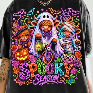 Może przedstawiać: Czarny t-shirt z neonową grafiką przedstawiającą trzy krowy rasy Highland przebrane na Halloween. Jedna ma kapelusz czarownicy, druga jest duchem na rowerze, a trzecia mumią. Wz&oacute;r zawiera dynie, nietoperze, pajęczyny oraz napis SPOOKY SEASON.