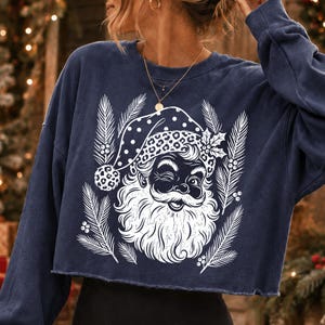 Puede incluir: Una sudadera corta de color azul marino con mangas largas y un estampado gr&aacute;fico blanco de un Santa Claus gui&ntilde;ando un ojo con un gorro de estampado de leopardo, rodeado de ramas de pino y bayas de acebo, con cuello redondo.