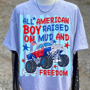 K&ouml;nnte beinhalten: Hellgraues T-Shirt mit einem Monster-Truck-Motiv in Rot, Wei&szlig; und Blau. Der Text lautet "ALL AMERICAN BOY RAISED ON MUD AND FREEDOM". Das Design umfasst Sterne und eine amerikanische Flagge.