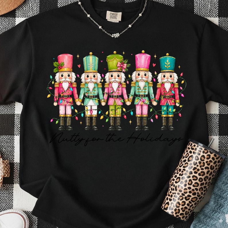 Op de afbeelding: Een zwart t-shirt met een afbeelding van vijf notenkrakers in roze, groene en turquoise uniformen die elkaars hand vasthouden, versierd met kleurrijke kerstverlichting en kleine rode hartjes. De tekst "Nutty for the Holidays" staat in het zwart eronder.