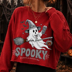 May include: A red long-sleeve cropped sweatshirt featuring a graphic of a ghost wearing a witch hat, riding a broomstick, holding a drink, and carrying a jack-o'-lantern. The text reads SPOOKY and HAPPY HAUNTING with spider web accents.