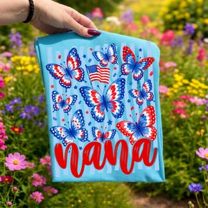 May include: A light blue t-shirt featuring a graphic print of multiple butterflies with red, white, and blue wing patterns, a small American flag, and the word "nana" written in large, bold red script lettering across the bottom section.