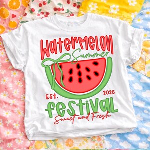 以下が含まれることがあります： スイカのスライスグラフィックがプリントされた白いTシャツ。「Watermelon Summer Festival」の文字が赤と緑でデザインされ、年号「2026」と「Sweet and Fresh」のフレーズが添えられています。